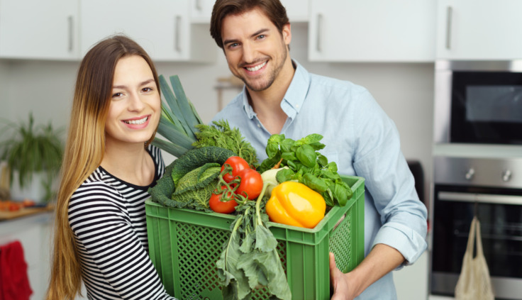 Ein junger Mann und eine junge Frau zu, die mit einem Lächeln eine volle Kiste mit frischem Gemüse tragen.