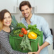 Ein junger Mann und eine junge Frau zu, die mit einem Lächeln eine volle Kiste mit frischem Gemüse tragen.
