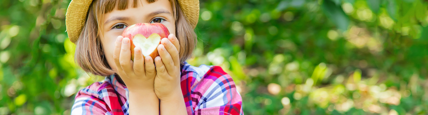 Auf dem Bild ist ein Mädchen mit einem glücklichen Lächeln zu sehen. Sie hält einen angebissenen Apfel in der Hand, dessen Biss eine Herzform ergibt. Bio Kinderverpflegung.