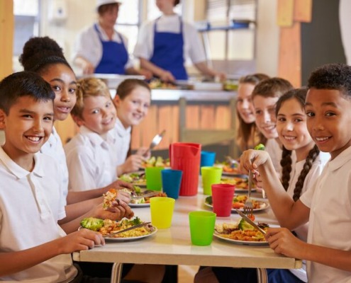 Schulcatering: Kinder sitzen am Tisch und genießen gesunde Kindermenüs.