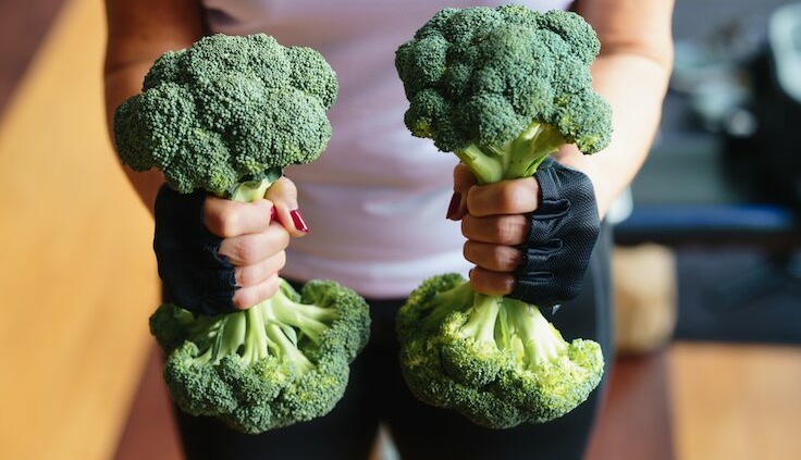 Eine Frau hält zwei Brokkoli in den Händen, die wie Fitnessstangen aussehen.