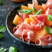 Honigmelone mit Parmaschinken und Basilikum. Das Gericht ist mit grobem Meersalz und frisch gemahlenem schwarzen Pfeffer gewürzt.