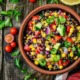 Quinoa-Salat mit Quinoa, Avocado, schwarzen Bohnen, Mais, roter Paprika, roten Zwiebeln, Jalapeño, Kirschtomaten und Petersilie. Im Hintergrund sind Kirschtomaten und Petersilienblätter zu sehen.