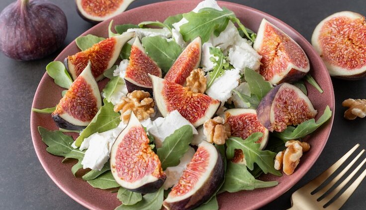 Ein frischer Salat aus reife Feigen, Rucola, Ziegenkäse und Walnüssen auf einem rosa Teller. Die süßen Früchte der Feige sind in Viertel geschnitten und kunstvoll auf dem Salat angerichtet. Im Hintergrund sind weitere geschnittene und ganze Feigen zu sehen, was die frischen Zutaten und die gesunde Ernährung hervorhebt. Eine goldene Gabel liegt bereit, um den leckeren und nährstoffreichen Snack zu genießen.