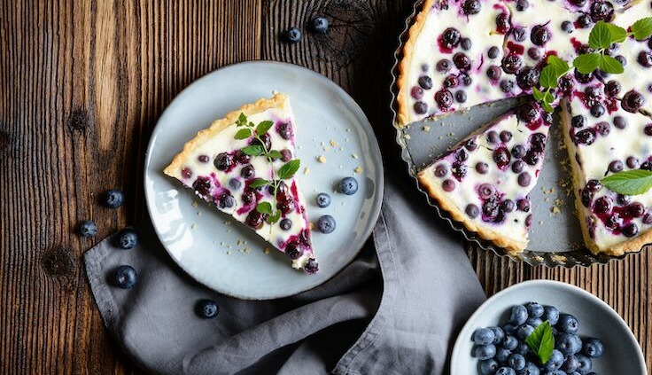 Ein köstliches Stück Heidelbeer-Torte liegt auf einem grauen Teller, dekoriert mit frischen Heidelbeeren und einem kleinen Minzblatt. Die restliche Torte befindet sich in einer Tarteform und ist ebenfalls mit vielen Heidelbeeren belegt.