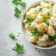 Ein frischer Kartoffelsalat mit cremiger Mayonnaise, garniert mit gehackter Petersilie und rosa Pfefferkörnern in einer weißen Schale, umgeben von dekorativen Kräutern auf einer hellen Unterlage.