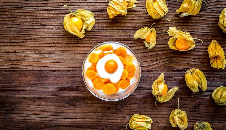 Eine Schale mit frischen, aufgeschnittenen Physalis-Früchten, die in Joghurt serviert werden. Um die Schale herum sind mehrere physalisfrüchte mit ihrer charakteristischen papierartigen Hülle auf einem Holztisch verteilt.