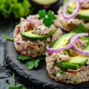 Das Gericht besteht aus Reispatties, die mit zerkleinertem Thunfisch, frischen Avocadoscheiben, roten Zwiebelringen und Petersilie garniert sind. Die lebendigen Farben und Texturen heben eine nahrhafte Mahlzeit hervor, die sich perfekt für eine ausgewogene Ernährung eignet.