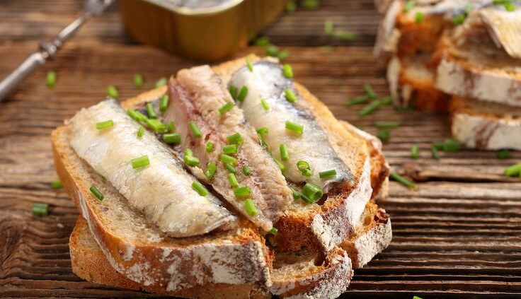 Sardinenfilets auf frischem Vollkornbrot, garniert mit gehacktem Schnittlauch, neben einer geöffneten Dose Sardinen serviert.