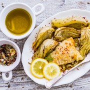 Gerösteter Fenchel mit gebratenem Fischfilet, serviert auf einem weißen Teller, garniert mit frischen Zitronenscheiben und einer Gewürzmischung. Im Hintergrund befinden sich Schälchen mit Olivenöl und Gewürzen.