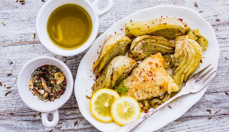 Gerösteter Fenchel mit gebratenem Fischfilet, serviert auf einem weißen Teller, garniert mit frischen Zitronenscheiben und einer Gewürzmischung. Im Hintergrund befinden sich Schälchen mit Olivenöl und Gewürzen.