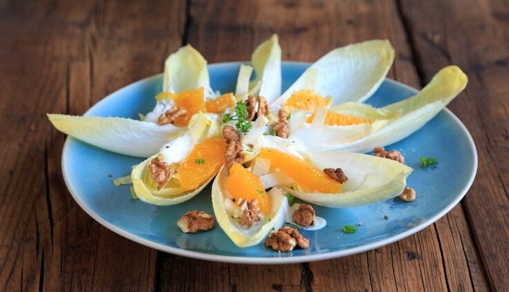 Salat aus Chicorée mit Orangenstücken, Walnüssen und frischem Dressing auf einem blauen Teller, angerichtet auf einem rustikalen Holztisch