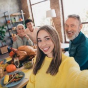 Junge Frau im gelben Pullover macht ein Selfie bei einem Familienessen. Im Hintergrund sitzen lächelnde Familienmitglieder an einem festlich gedeckten Tisch mit Truthahn, Gemüse, Kürbissen und Kerzen.