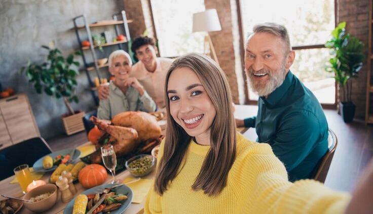 Junge Frau im gelben Pullover macht ein Selfie bei einem Familienessen. Im Hintergrund sitzen lächelnde Familienmitglieder an einem festlich gedeckten Tisch mit Truthahn, Gemüse, Kürbissen und Kerzen.