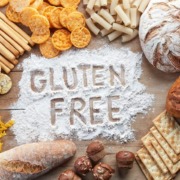 Verschiedene glutenfreie Lebensmittel wie Brot, Cracker, Pasta und Snacks, arrangiert um die Worte 'Gluten Free', die in Mehl auf einer Holzoberfläche geschrieben sind.