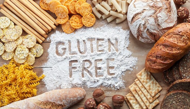 Verschiedene glutenfreie Lebensmittel wie Brot, Cracker, Pasta und Snacks, arrangiert um die Worte 'Gluten Free', die in Mehl auf einer Holzoberfläche geschrieben sind.