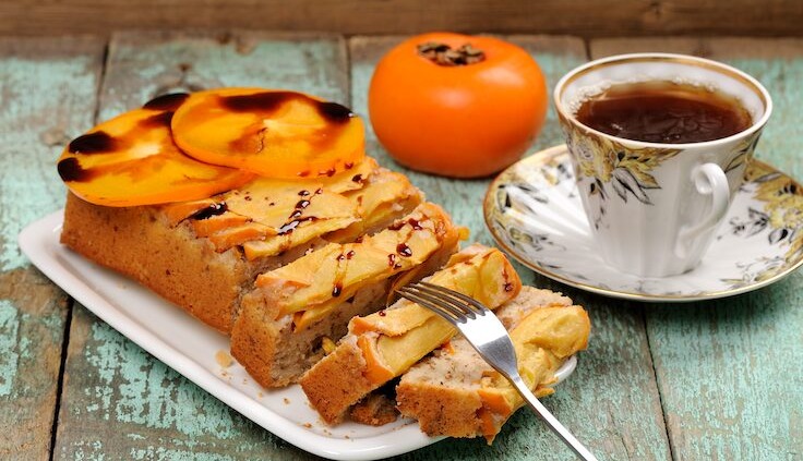 Saftiger Kuchen mit Kaki-Früchten, serviert auf einem weißen Teller mit einer Tasse Tee auf einem rustikalen Holztisch.