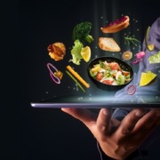 Hand hält ein Tablet mit einer digitalen Darstellung von frischen Zutaten und einem Salat, die scheinbar in der Luft schweben – symbolisiert modernes Online-Bestellen von gesundem Essen.