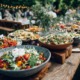 Rustikales Outdoor-Catering mit einer Auswahl an bunten Salaten in großen Schüsseln auf Holztischen, im Hintergrund Gäste in entspannter Gartenatmosphäre.