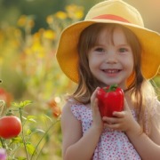 Lächelndes Mädchen mit gelbem Sonnenhut hält eine rote Paprika in der Hand, umgeben von blühenden Blumen und Tomatenpflanzen – Symbol für gesunde Ernährung und frisches Gemüse im Sommer.