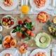 Gesundes Catering-Buffet mit einer Vielzahl bunter Früchte, Smoothies, Fruchtsäften und stilvoll angerichteten Tellern – dekorativ präsentiert auf einem hellen Holztisch mit frischen Erdbeeren, Kiwis, Papayas, Beeren, Äpfeln und exotischen Obstsorten.