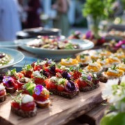 Rustikales Buffet mit gesunden, bunt angerichteten Häppchen auf Holzplatten, serviert im Freien mit Blumendeko im Hintergrund