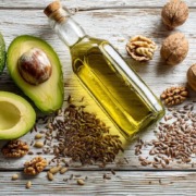 Ein wunderschön arrangiertes Flat-Lay gesunder Fette – darunter eine Flasche natives Olivenöl extra, reife Avocadohälften, Walnüsse, Leinsamen und verstreute Sonnenblumenkerne. Natürliches Licht, weiche Schatten, minimalistischer Holzuntergrund, hochauflösender Food-Stil.