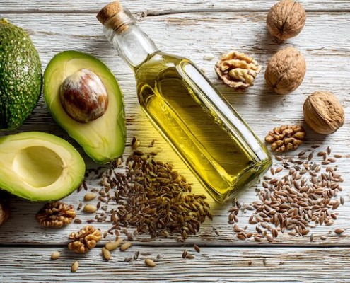 Ein wunderschön arrangiertes Flat-Lay gesunder Fette – darunter eine Flasche natives Olivenöl extra, reife Avocadohälften, Walnüsse, Leinsamen und verstreute Sonnenblumenkerne. Natürliches Licht, weiche Schatten, minimalistischer Holzuntergrund, hochauflösender Food-Stil.