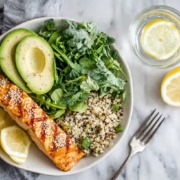 Gesundes Gericht mit gegrilltem Lachs, Avocado, Quinoa, frischem Blattsalat und Zitronenscheiben, serviert mit einem Glas Wasser mit Zitrone