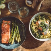 Zwei gesunde Lachsgerichte auf einem rustikalen Holztisch: links gegrillter Wildlachs mit grünem Spargel und Zitrone, rechts Tagliatelle mit Wildlachs, grünem Spargel und Kräuterpesto – stilvoll angerichtet, mit frischem Wasser und einer Zitrone im Hintergrund.