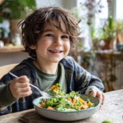 Ein fröhliches Kind sitzt am Esstisch und isst einen bunten Salat mit Feldsalat(Valerianella locusta), Karotten und Äpfeln, helle freundliche Atmosphäre, natürliche Farben, Bio-Lifestyle