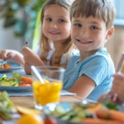 Zwei lächelnde Kinder essen gemeinsam ein gesundes Mittagessen mit frischem Gemüse und Saft am Tisch.