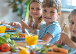 Zwei lächelnde Kinder essen gemeinsam ein gesundes Mittagessen mit frischem Gemüse und Saft am Tisch.