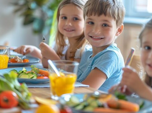 Zwei lächelnde Kinder essen gemeinsam ein gesundes Mittagessen mit frischem Gemüse und Saft am Tisch.