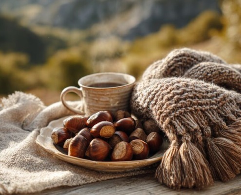 Eine gemütliche herbstliche Picknickszene mit gerösteten Maronen, einem warmen Getränk, einem gestrickten Schal, sanftem Sonnenlicht, Bergen oder Wald im Hintergrund, in natürlichen, warmen Farbtönen.