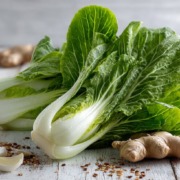 pak_choi_gesund Frisch angerichteter Pak Choi auf einem hellen Holztisch, mit glänzenden grünen Blättern und weißen Stielen, daneben frischer Ingwer, Knoblauch und Sesamsamen, natürliches Licht, modernes Food-Styling, asiatisches Flair