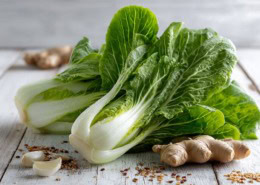 Frisch angerichteter Pak Choi auf einem hellen Holztisch, mit glänzenden grünen Blättern und weißen Stielen, daneben frischer Ingwer, Knoblauch und Sesamsamen, natürliches Licht, modernes Food-Styling, asiatisches Flair