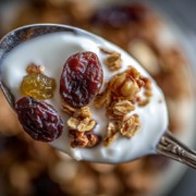 Nahaufnahme eines Löffels mit Müsli, Joghurt und Trockenpflaumen – die Trockenpflaumen stehen im Fokus, weich, glänzend und saftig. Daneben ein Glas mit warmer Milch und Bio-Honig. Natürliches Winterlicht, minimalistische Food-Fotografie auf rustikalem Holztisch, helle warme Holz- und Cremetöne, sanfter Unschärfeverlauf, gemütliche Atmosphäre