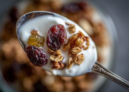 Nahaufnahme eines Löffels mit Müsli, Joghurt und Trockenpflaumen – die Trockenpflaumen stehen im Fokus, weich, glänzend und saftig. Daneben ein Glas mit warmer Milch und Bio-Honig. Natürliches Winterlicht, minimalistische Food-Fotografie auf rustikalem Holztisch, helle warme Holz- und Cremetöne, sanfter Unschärfeverlauf, gemütliche Atmosphäre