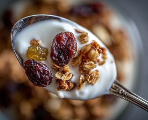 Nahaufnahme eines Löffels mit Müsli, Joghurt und Trockenpflaumen – die Trockenpflaumen stehen im Fokus, weich, glänzend und saftig. Daneben ein Glas mit warmer Milch und Bio-Honig. Natürliches Winterlicht, minimalistische Food-Fotografie auf rustikalem Holztisch, helle warme Holz- und Cremetöne, sanfter Unschärfeverlauf, gemütliche Atmosphäre
