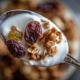 Nahaufnahme eines Löffels mit Müsli, Joghurt und Trockenpflaumen – die Trockenpflaumen stehen im Fokus, weich, glänzend und saftig. Daneben ein Glas mit warmer Milch und Bio-Honig. Natürliches Winterlicht, minimalistische Food-Fotografie auf rustikalem Holztisch, helle warme Holz- und Cremetöne, sanfter Unschärfeverlauf, gemütliche Atmosphäre