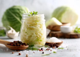 Schön arrangiertes Glas mit fermentiertem Sauerkraut, umgeben von Weißkohl, Salz und Gewürzen; sauberes, helles natürliches Licht, minimalistischer Wellness-Stil, probiotisches Lebensmittel.