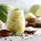 Schön arrangiertes Glas mit fermentiertem Sauerkraut, umgeben von Weißkohl, Salz und Gewürzen; sauberes, helles natürliches Licht, minimalistischer Wellness-Stil, probiotisches Lebensmittel.