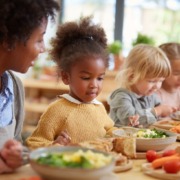 kindergarten mealtime, children eating together calmly, educator sitting nearby, supportive atmosphere, focus on community and learning, healthy food on table, vegetables, soup, bread,