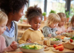 kindergarten mealtime, children eating together calmly, educator sitting nearby, supportive atmosphere, focus on community and learning, healthy food on table, vegetables, soup, bread,
