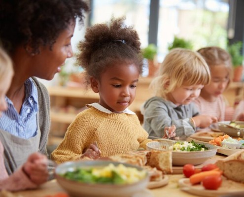 kindergarten mealtime, children eating together calmly, educator sitting nearby, supportive atmosphere, focus on community and learning, healthy food on table, vegetables, soup, bread,