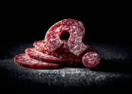 Nahaufnahme von Salami-Scheiben in Form eines Fragezeichens auf dunklem Hintergrund, dramatisches Spotlicht, hoher Kontrast, künstlerische Food-Fotografie mit geringer Tiefenschärfe.