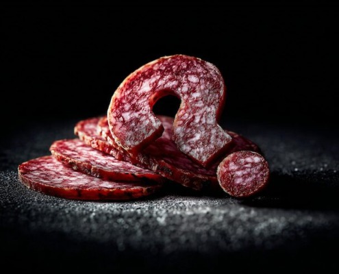 Nahaufnahme von Salami-Scheiben in Form eines Fragezeichens auf dunklem Hintergrund, dramatisches Spotlicht, hoher Kontrast, künstlerische Food-Fotografie mit geringer Tiefenschärfe.