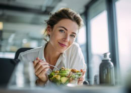 Lächelnde Geschäftsfrau schaut in die Kamera und isst eine gesunde Lunch-Bowl mit Gemüse, Avocado und Quinoa während ihrer Mittagspause im Büro.