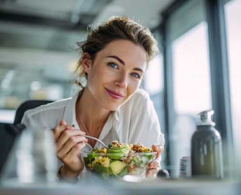 Lächelnde Geschäftsfrau schaut in die Kamera und isst eine gesunde Lunch-Bowl mit Gemüse, Avocado und Quinoa während ihrer Mittagspause im Büro.
